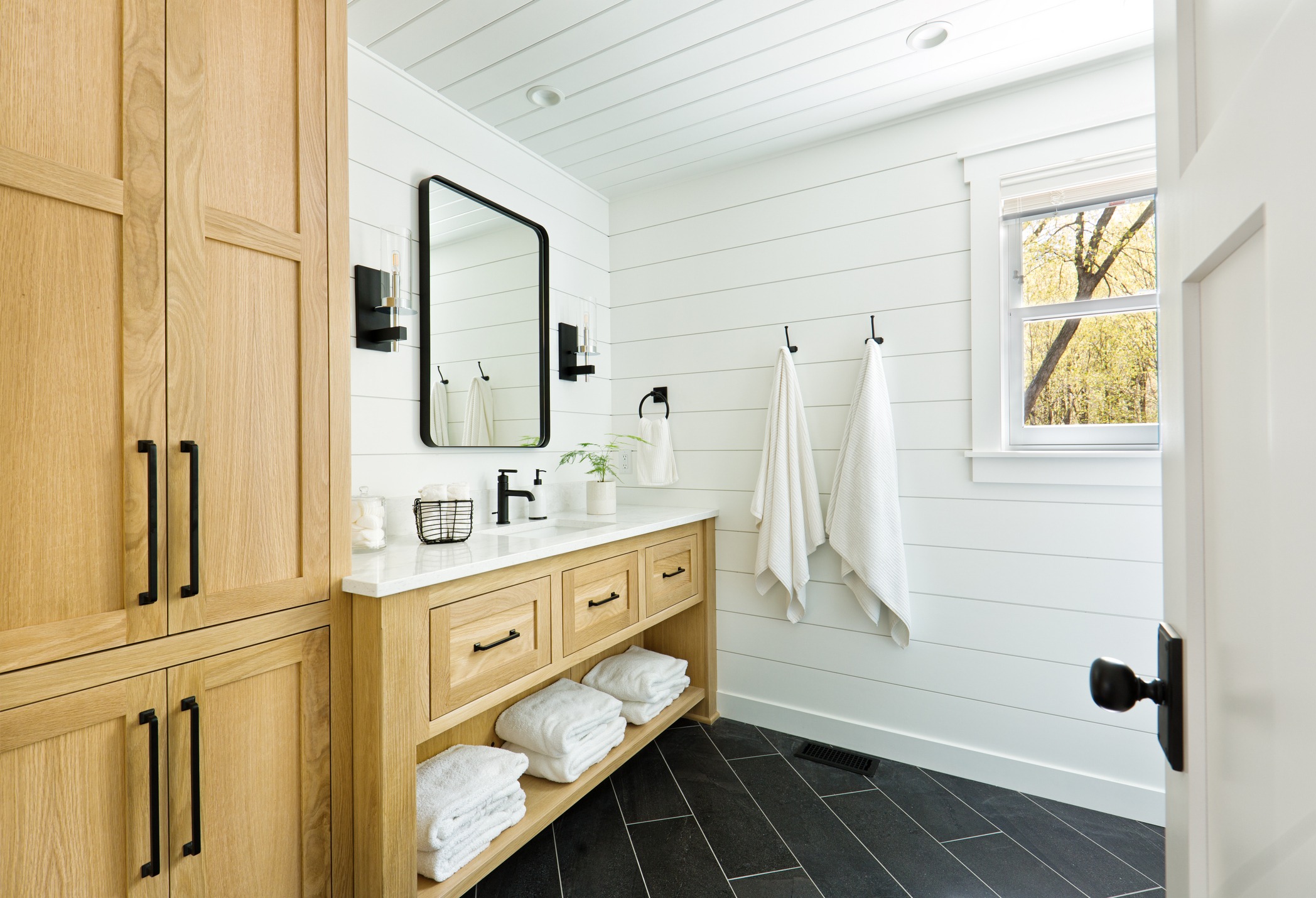 A newly renovated bathroom combines modern design with a contemporary country style with unfinished wooden cabinets and black tile.
